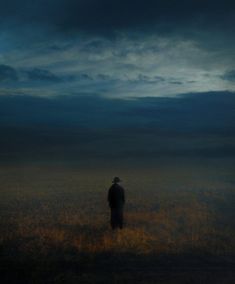 a lone man standing in the middle of a field under a dark sky with clouds