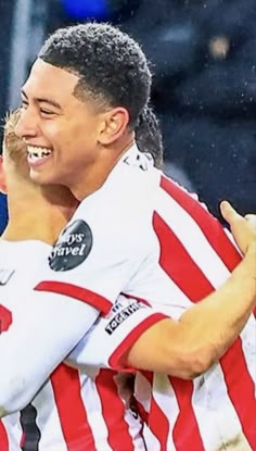 two soccer players are hugging each other on the field in front of an audience and one is wearing a red and white striped shirt