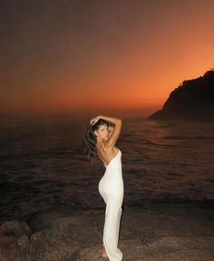 a woman standing on top of a rocky beach next to the ocean at sunset with her arms behind her head