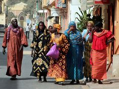 four women walking down the street in different colored dresses and head coverings, one carrying a purple bag