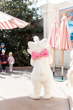 a large white teddy bear with a pink bow on it's neck standing in front of some umbrellas