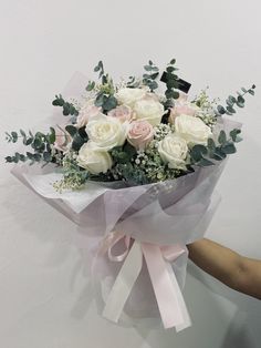 a bouquet of white roses and greenery wrapped in pink ribbon with someone's hand holding it