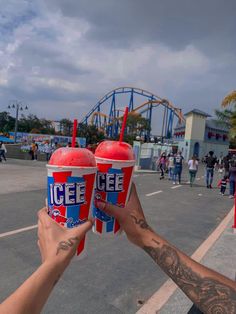 two people holding up red and blue drinks in front of an amusement park roller coaster