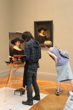 two people standing in front of an easel with paintings on the wall behind them