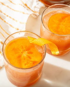 two glasses filled with orange juice sitting on top of a white tablecloth next to an orange slice