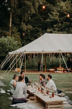 a group of people sitting around a wooden table in front of a tent with lights