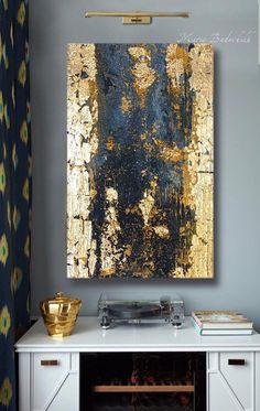 an abstract painting hangs on the wall above a white cabinet with drawers and a gold bowl