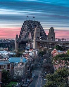 the sydney harbour bridge is shown in this view from across the street at sunset, with cars parked on the side of the road