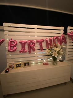 a birthday sign with pink balloons and flowers on a white table in front of a wall