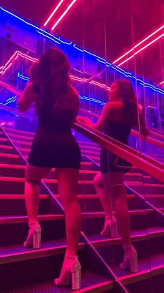 two women in short skirts are walking down stairs with neon lights behind them and one is wearing high heels