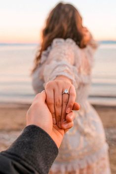 a man and woman holding hands while standing on the beach at sunset with their engagement ring in hand