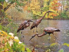 three flamingos are standing in the water near some trees and bushes with their legs spread out