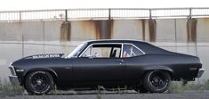 an old black car is parked in front of a rusty wall and grass near the road