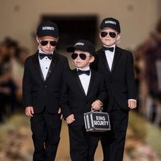 three little boys dressed up in black suits and hats, one holding a bag while the other is wearing sunglasses