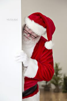 a man dressed as santa claus peeking out from behind a white board with his hands on it