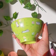 a hand holding a green bowl with cats on it and flowers in the background,