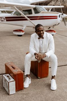 a man in a white suit and bow tie sitting on suitcases next to an airplane