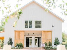 a large white barn with wooden shutters and windows on the front door is surrounded by greenery