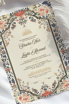 an ornate wedding card with flowers and leaves on it, sitting next to a feather