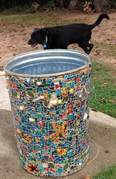 a black dog standing on top of a metal trash can covered in colorful glass tiles