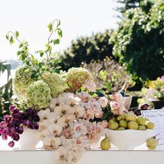 an arrangement of flowers and fruit in bowls on a white table with a sign that says,