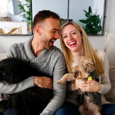 a man and woman sitting on a couch with two dogs in their lap, smiling at the camera