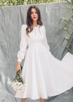 a woman in a white dress is holding a bouquet and standing next to a wall