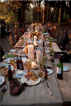 a long table is set up with candles and plates for an outdoor dinner party in the woods