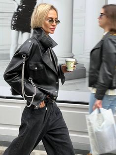 a woman walking down the street with a cup in her hand and wearing black leather jacket