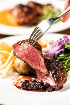 a person is holding a fork over some meat and potatoes on a white plate with other food