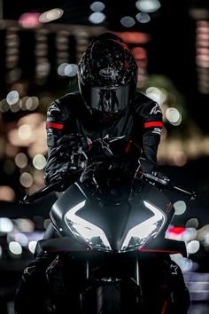 a man riding on the back of a black motorcycle at night with lights in the background