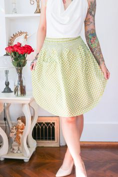 a woman standing in front of a shelf with flowers and tattoos on her arm, wearing a white top and green checkered skirt