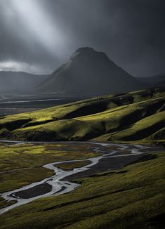 a river running through a lush green valley under a dark sky with mountains in the background