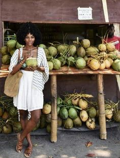 a woman standing in front of a fruit stand with coconuts and other tropical fruits