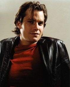 a man in a red shirt and black jacket looking at the camera while wearing a leather jacket