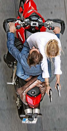 two people are sitting on a motorcycle looking down at the camera man and woman standing next to each other