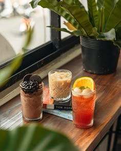 three drinks are sitting on a table next to a book and potted plant in front of a window