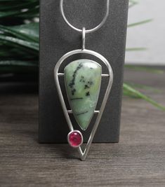 Meadows Pendant A unique specimen of chrysocolla is showcased in this sleek modern themed pendant.   What makes the stone unique are the  inclusions of plume agate.   The lovely minty color of this beautifully cut and polished stone is accented by a hot pink tourmaline gemstone. I set both of the stones in fine silver with solid silver backs.  They are connected to a framework of thick sterling wire that has a round profile. Overall this pendant measures about 1-1/4" wide x 2-3/4" long (includin Modern Silver Jewelry Pendant, Silversmith Pendant, Round Profile, Modern Silver Jewelry, Plume Agate, Unique Pendant Necklace, Wire Jewelry Designs, Silver Jewelry Design, Design Statement