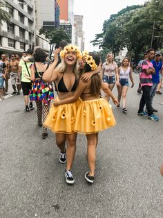 two women in yellow skirts are walking down the street with other people behind them and one woman is holding her head up