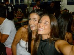 two young women sitting next to each other at a table with people in the background