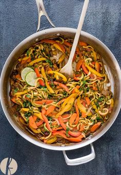 a pan filled with noodles and vegetables on top of a blue table cloth next to a wooden spoon