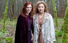 two women standing next to each other in front of green moss covered trees and bushes