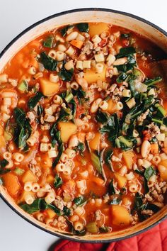 a pot filled with pasta and spinach soup on top of a red cloth next to a wooden spoon