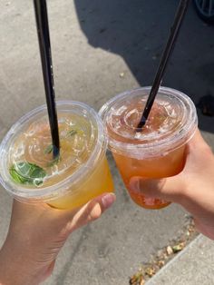 two people holding drinks with straws in their hands on the sidewalk, one is orange and the other is lemonade