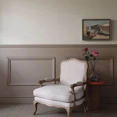 a white chair sitting in front of a painting on the wall next to a vase with flowers