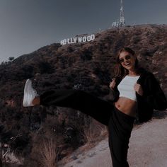 a woman standing on top of a dirt road next to a hill with the hollywood sign in the background