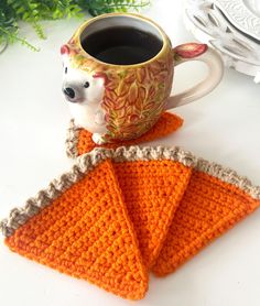 an orange crocheted napkin with a coffee cup on it and a mug in the background