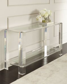 a clear console table with flowers on top in a room next to a white rug