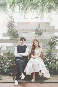 two people sitting on chairs in front of flowers and greenery, one person is wearing a white dress