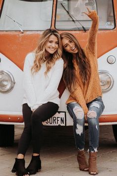 two women sitting on the side of a bus posing for a photo together with their arms in the air